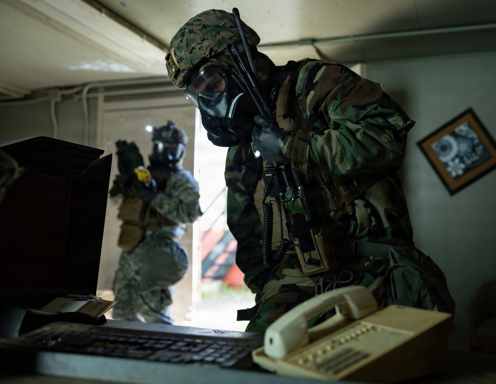 Two military personnel wearing full CBRN (Chemical, Biological, Radiological, Nuclear) protective gear including gas masks and combat uniforms are conducting what appears to be a search or clearance operation inside a building. The foreground soldier, wearing woodland camouflage and a MARPAT helmet, is leaning over a desk examining a computer workstation. A second soldier in digital camouflage stands near a doorway in the background.