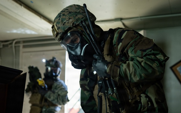 Two military personnel wearing full CBRN (Chemical, Biological, Radiological, Nuclear) protective gear including gas masks and combat uniforms are conducting what appears to be a search or clearance operation inside a building. The foreground soldier, wearing woodland camouflage and a MARPAT helmet, is leaning over a desk examining a computer workstation. A second soldier in digital camouflage stands near a doorway in the background.