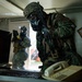 Two military personnel wearing full CBRN (Chemical, Biological, Radiological, Nuclear) protective gear including gas masks and combat uniforms are conducting what appears to be a search or clearance operation inside a building. The foreground soldier, wearing woodland camouflage and a MARPAT helmet, is leaning over a desk examining a computer workstation. A second soldier in digital camouflage stands near a doorway in the background.