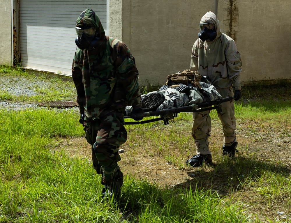 Two military personnel wearing CBRN (Chemical, Biological, Radiological, Nuclear) protective suits and gas masks are carrying a person on a stretcher across a grassy area near a building. One individual is wearing woodland camouflage pattern while the other wears desert camouflage (DCU/3-color desert pattern). This appears to be a military training exercise involving casualty evacuation under CBRN conditions.