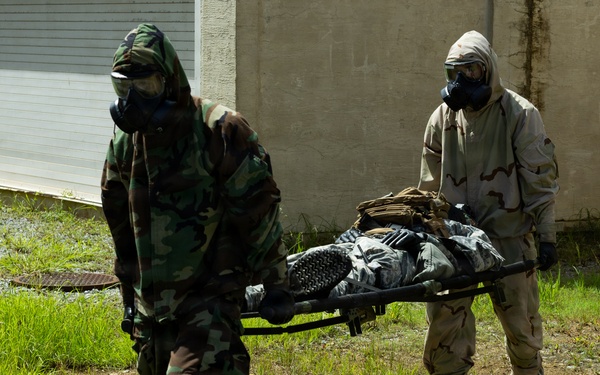 Two military personnel wearing CBRN (Chemical, Biological, Radiological, Nuclear) protective suits and gas masks are carrying a person on a stretcher across a grassy area near a building. One individual is wearing woodland camouflage pattern while the other wears desert camouflage (DCU/3-color desert pattern). This appears to be a military training exercise involving casualty evacuation under CBRN conditions.