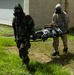 Two military personnel wearing CBRN (Chemical, Biological, Radiological, Nuclear) protective suits and gas masks are carrying a person on a stretcher across a grassy area near a building. One individual is wearing woodland camouflage pattern while the other wears desert camouflage (DCU/3-color desert pattern). This appears to be a military training exercise involving casualty evacuation under CBRN conditions.