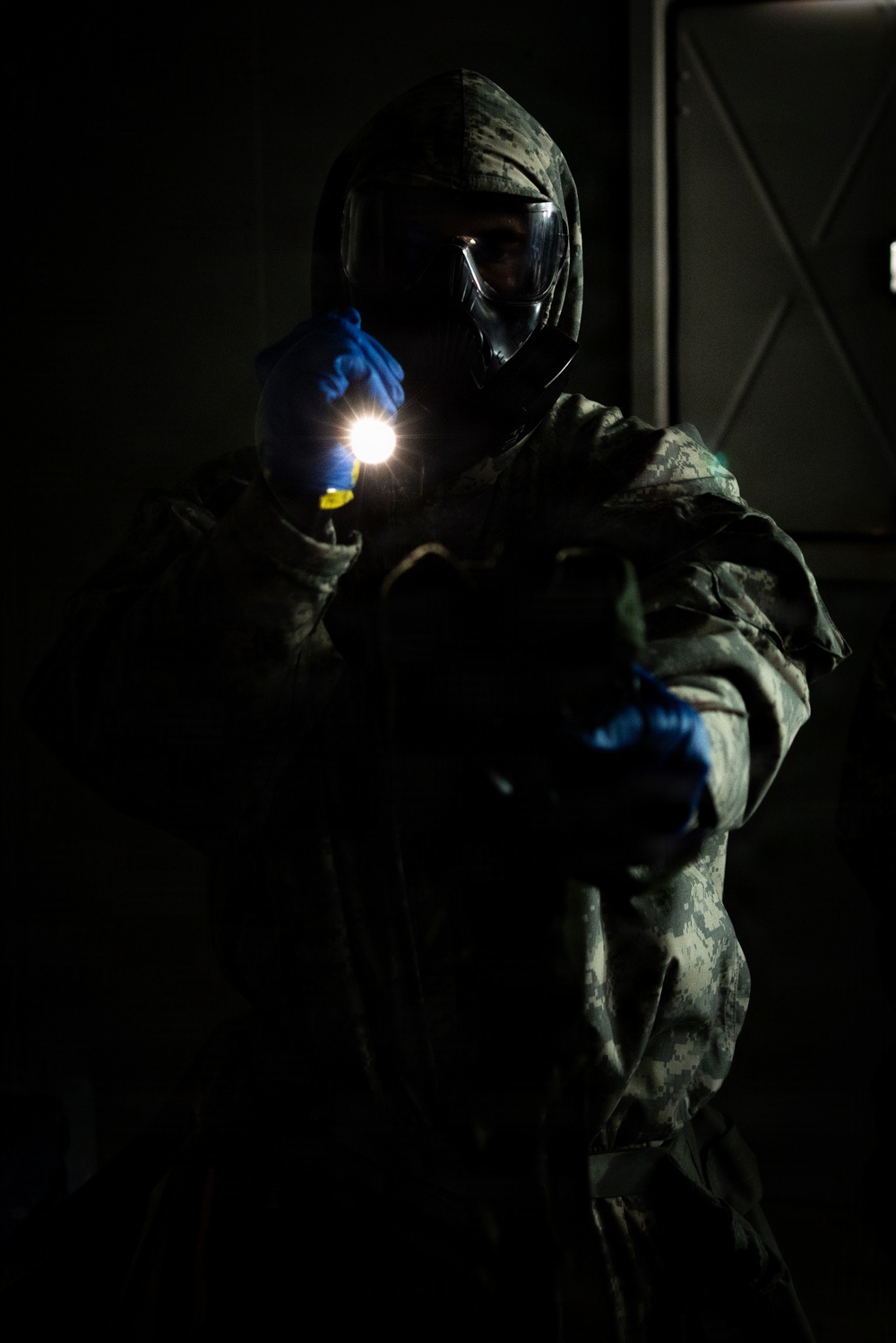 A person wearing full CBRN (Chemical, Biological, Radiological, Nuclear) protective gear, including a gas mask, digital camouflage uniform, and blue nitrile gloves, holds an illuminated flashlight in a dark environment. The dramatic low-key lighting highlights the figure against a nearly black background. The setting appears to be an indoor or enclosed space, possibly during a training exercise or operational scenario.