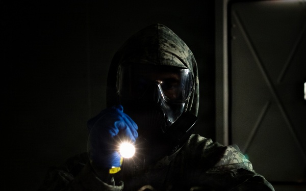 A person wearing full CBRN (Chemical, Biological, Radiological, Nuclear) protective gear, including a gas mask, digital camouflage uniform, and blue nitrile gloves, holds an illuminated flashlight in a dark environment. The dramatic low-key lighting highlights the figure against a nearly black background. The setting appears to be an indoor or enclosed space, possibly during a training exercise or operational scenario.