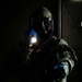 A person wearing full CBRN (Chemical, Biological, Radiological, Nuclear) protective gear, including a gas mask, digital camouflage uniform, and blue nitrile gloves, holds an illuminated flashlight in a dark environment. The dramatic low-key lighting highlights the figure against a nearly black background. The setting appears to be an indoor or enclosed space, possibly during a training exercise or operational scenario.