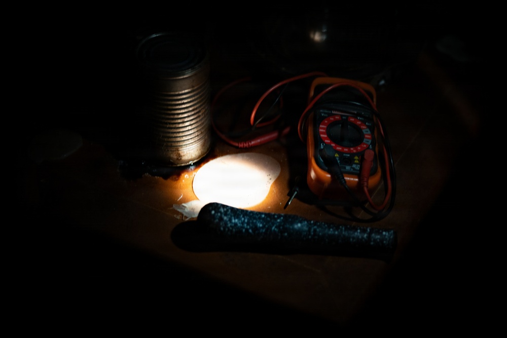A dimly lit workspace showing a collection of electronic testing and hand tools arranged on a wooden surface. A bright light source illuminates the center of the scene, casting dramatic shadows. The items appear to be part of an informal electronics or DIY repair setup.