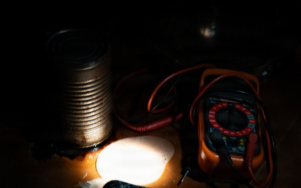 A dimly lit workspace showing a collection of electronic testing and hand tools arranged on a wooden surface. A bright light source illuminates the center of the scene, casting dramatic shadows. The items appear to be part of an informal electronics or DIY repair setup.