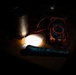 A dimly lit workspace showing a collection of electronic testing and hand tools arranged on a wooden surface. A bright light source illuminates the center of the scene, casting dramatic shadows. The items appear to be part of an informal electronics or DIY repair setup.