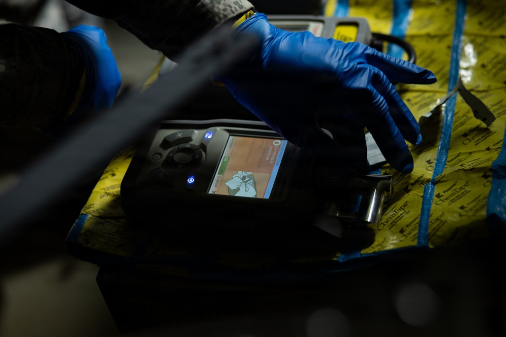 A person wearing blue nitrile gloves is operating a handheld electronic detection or analysis device, likely a chemical or gas detector, placed on a yellow surface secured with blue tape. The device has an illuminated color screen displaying what appears to be a warning or instructional graphic. The setting appears to be a field or industrial environment, suggesting a hazmat, forensic, or safety inspection context.