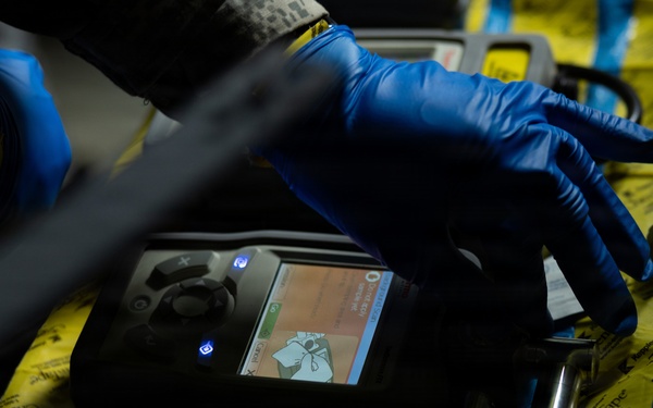 A person wearing blue nitrile gloves is operating a handheld electronic detection or analysis device, likely a chemical or gas detector, placed on a yellow surface secured with blue tape. The device has an illuminated color screen displaying what appears to be a warning or instructional graphic. The setting appears to be a field or industrial environment, suggesting a hazmat, forensic, or safety inspection context.