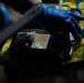 A person wearing blue nitrile gloves is operating a handheld electronic detection or analysis device, likely a chemical or gas detector, placed on a yellow surface secured with blue tape. The device has an illuminated color screen displaying what appears to be a warning or instructional graphic. The setting appears to be a field or industrial environment, suggesting a hazmat, forensic, or safety inspection context.