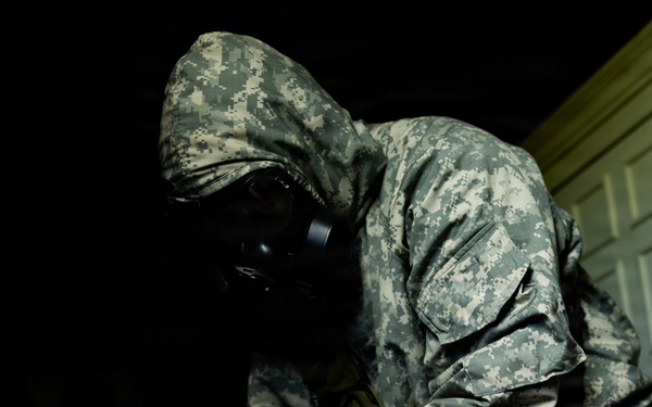 A person wearing US Army ACU (Army Combat Uniform) digital camouflage pattern is equipped with a full-face gas mask and blue nitrile gloves, handling a small object or instrument in a low-light environment. The individual appears to be conducting a CBRN (Chemical, Biological, Radiological, Nuclear) response or hazmat operation based on the protective equipment worn. A partially visible plastic bag with contents is present in the lower right of the frame.