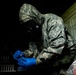 A person wearing US Army ACU (Army Combat Uniform) digital camouflage pattern is equipped with a full-face gas mask and blue nitrile gloves, handling a small object or instrument in a low-light environment. The individual appears to be conducting a CBRN (Chemical, Biological, Radiological, Nuclear) response or hazmat operation based on the protective equipment worn. A partially visible plastic bag with contents is present in the lower right of the frame.