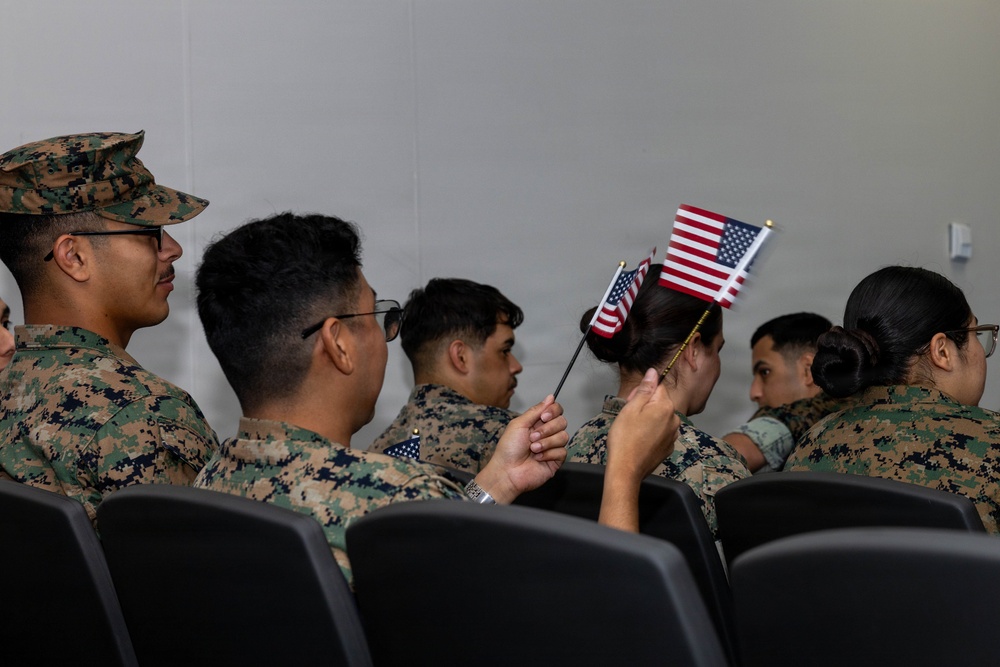 A group of individuals wearing U.S. Marine Corps MARPAT (digital camouflage) uniforms are seated in rows of black chairs in what appears to be an indoor ceremony or briefing room. One person in the center is holding two small American flags, suggesting this may be a naturalization ceremony or patriotic military event. The scene captures a formal yet intimate moment among service members.