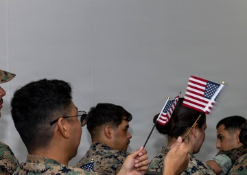 A group of individuals wearing U.S. Marine Corps MARPAT (digital camouflage) uniforms are seated in rows of black chairs in what appears to be an indoor ceremony or briefing room. One person in the center is holding two small American flags, suggesting this may be a naturalization ceremony or patriotic military event. The scene captures a formal yet intimate moment among service members.
