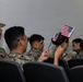 A group of individuals wearing U.S. Marine Corps MARPAT (digital camouflage) uniforms are seated in rows of black chairs in what appears to be an indoor ceremony or briefing room. One person in the center is holding two small American flags, suggesting this may be a naturalization ceremony or patriotic military event. The scene captures a formal yet intimate moment among service members.