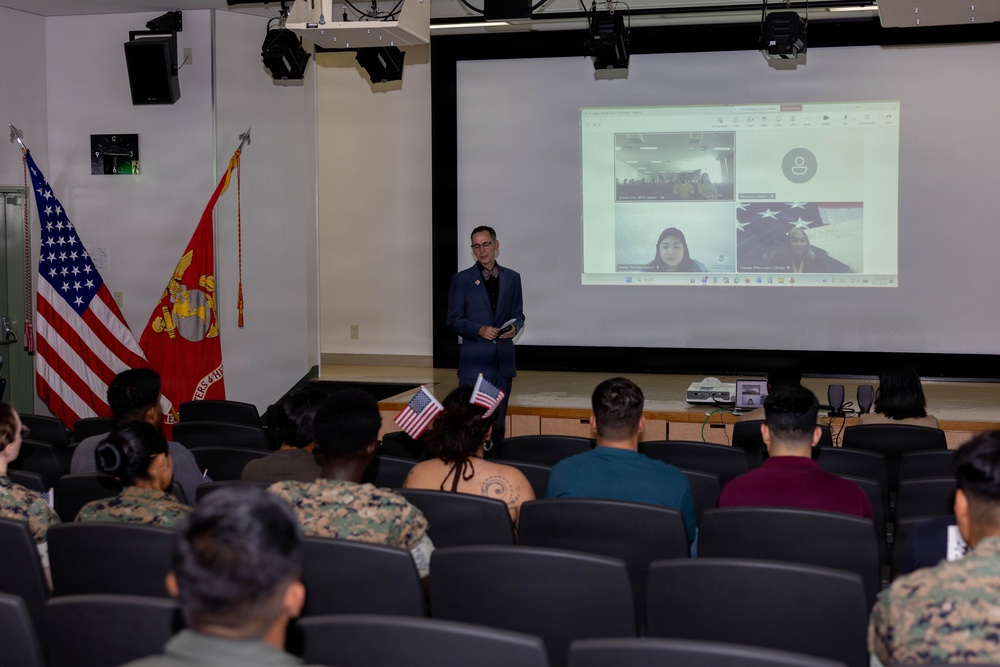 A formal presentation or ceremony is taking place in a military auditorium, with a civilian speaker standing before an audience that includes uniformed Marines and civilians. A video conference is being projected on a large screen behind the speaker, showing remote participants. The presence of American flags, a United States Marine Corps flag, and small handheld American flags suggest this may be a naturalization ceremony or patriotic event.