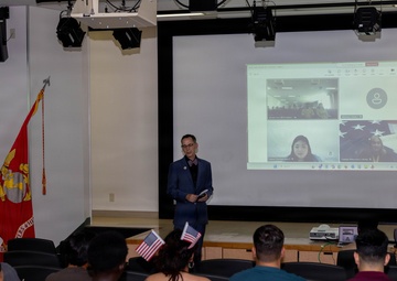 A formal presentation or ceremony is taking place in a military auditorium, with a civilian speaker standing before an audience that includes uniformed Marines and civilians. A video conference is being projected on a large screen behind the speaker, showing remote participants. The presence of American flags, a United States Marine Corps flag, and small handheld American flags suggest this may be a naturalization ceremony or patriotic event.