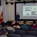A formal presentation or ceremony is taking place in a military auditorium, with a civilian speaker standing before an audience that includes uniformed Marines and civilians. A video conference is being projected on a large screen behind the speaker, showing remote participants. The presence of American flags, a United States Marine Corps flag, and small handheld American flags suggest this may be a naturalization ceremony or patriotic event.