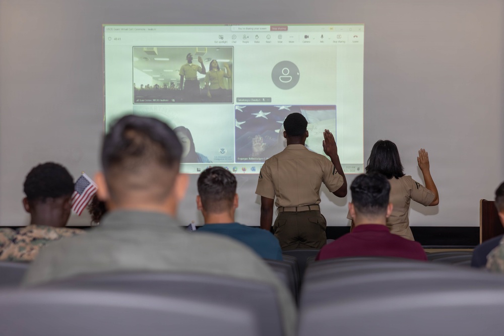 A virtual ceremony is taking place in what appears to be a military setting, where two individuals in uniform are raising their right hands facing a projected screen showing a video conference. The projected screen displays participants also raising their hands, suggesting this is a naturalization or oath-taking ceremony conducted remotely. Several seated attendees, some in military camouflage uniforms, observe the proceedings, and a small American flag is visible among them.