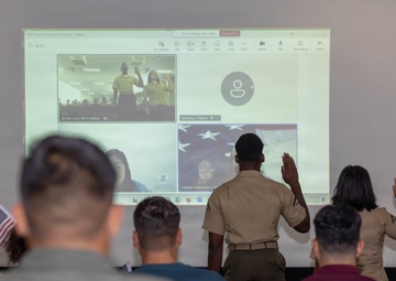 A virtual ceremony is taking place in what appears to be a military setting, where two individuals in uniform are raising their right hands facing a projected screen showing a video conference. The projected screen displays participants also raising their hands, suggesting this is a naturalization or oath-taking ceremony conducted remotely. Several seated attendees, some in military camouflage uniforms, observe the proceedings, and a small American flag is visible among them.