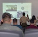 A virtual ceremony is taking place in what appears to be a military setting, where two individuals in uniform are raising their right hands facing a projected screen showing a video conference. The projected screen displays participants also raising their hands, suggesting this is a naturalization or oath-taking ceremony conducted remotely. Several seated attendees, some in military camouflage uniforms, observe the proceedings, and a small American flag is visible among them.