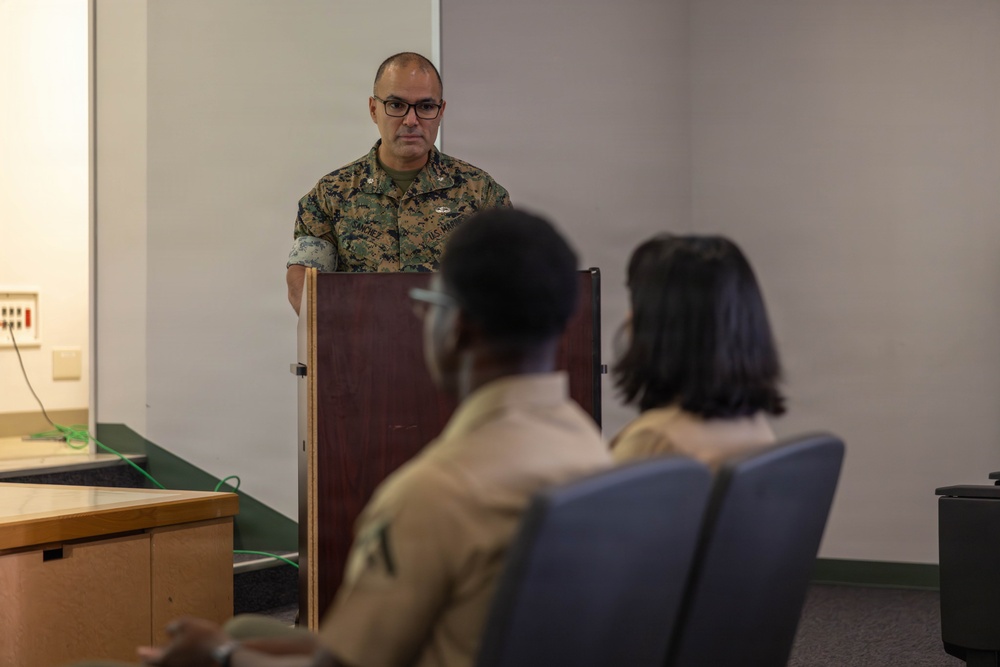 A U.S. Marine in woodland MARPAT camouflage uniform stands at a wooden podium, appearing to address or brief a small audience. Two individuals are seated in the foreground with their backs to the camera, listening to the speaker. The setting appears to be a briefing room or classroom within a military facility.