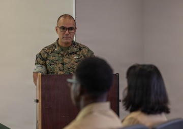 A U.S. Marine in woodland MARPAT camouflage uniform stands at a wooden podium, appearing to address or brief a small audience. Two individuals are seated in the foreground with their backs to the camera, listening to the speaker. The setting appears to be a briefing room or classroom within a military facility.