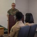 A U.S. Marine in woodland MARPAT camouflage uniform stands at a wooden podium, appearing to address or brief a small audience. Two individuals are seated in the foreground with their backs to the camera, listening to the speaker. The setting appears to be a briefing room or classroom within a military facility.