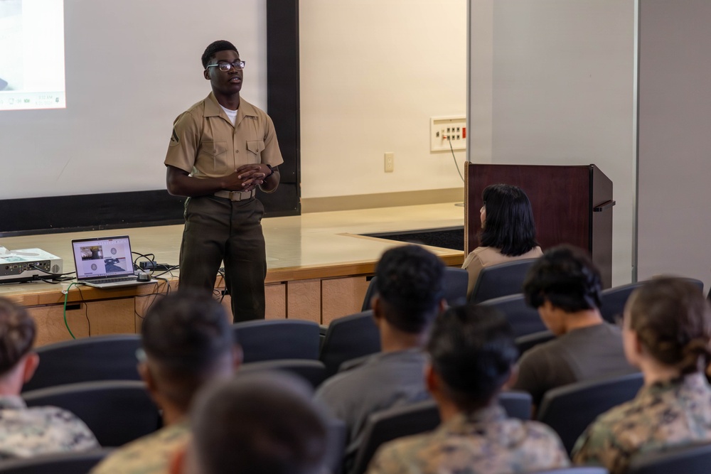 A military service member in tan and olive green uniform is delivering a presentation to an audience of fellow service members in a briefing room or auditorium. A laptop and projector are set up on a table near the stage, displaying content on a screen behind the presenter. The audience, wearing various military uniforms including camouflage patterns, is seated in rows of chairs facing the presenter.
