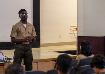 A military service member in tan and olive green uniform is delivering a presentation to an audience of fellow service members in a briefing room or auditorium. A laptop and projector are set up on a table near the stage, displaying content on a screen behind the presenter. The audience, wearing various military uniforms including camouflage patterns, is seated in rows of chairs facing the presenter.