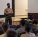 A military service member in tan and olive green uniform is delivering a presentation to an audience of fellow service members in a briefing room or auditorium. A laptop and projector are set up on a table near the stage, displaying content on a screen behind the presenter. The audience, wearing various military uniforms including camouflage patterns, is seated in rows of chairs facing the presenter.