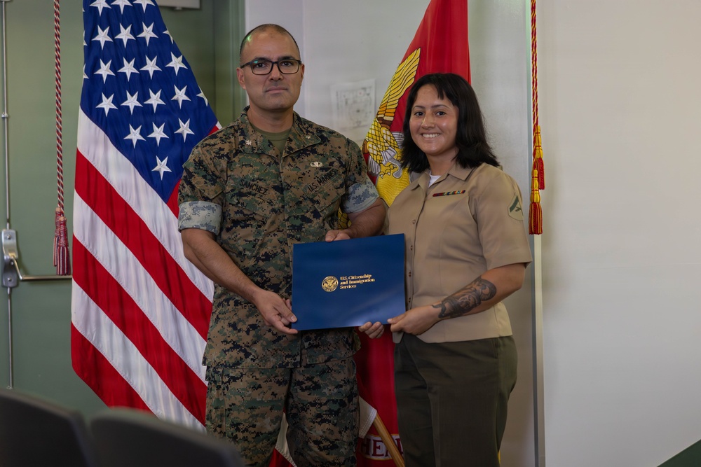 Two U.S. Marines pose together holding a blue folder bearing the U.S. Citizenship and Immigration Services (USCIS) seal, suggesting a naturalization ceremony. The male Marine on the left wears camouflage MARPAT utilities with the name tape 'Sanchez,' while the female Marine on the right wears the tan Service 'C' uniform with ribbons and a Lance Corporal chevron. They are standing in front of a U.S. flag and a Marine Corps flag.