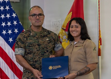 Two U.S. Marines pose together holding a blue folder bearing the U.S. Citizenship and Immigration Services (USCIS) seal, suggesting a naturalization ceremony. The male Marine on the left wears camouflage MARPAT utilities with the name tape 'Sanchez,' while the female Marine on the right wears the tan Service 'C' uniform with ribbons and a Lance Corporal chevron. They are standing in front of a U.S. flag and a Marine Corps flag.