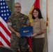 Two U.S. Marines pose together holding a blue folder bearing the U.S. Citizenship and Immigration Services (USCIS) seal, suggesting a naturalization ceremony. The male Marine on the left wears camouflage MARPAT utilities with the name tape 'Sanchez,' while the female Marine on the right wears the tan Service 'C' uniform with ribbons and a Lance Corporal chevron. They are standing in front of a U.S. flag and a Marine Corps flag.