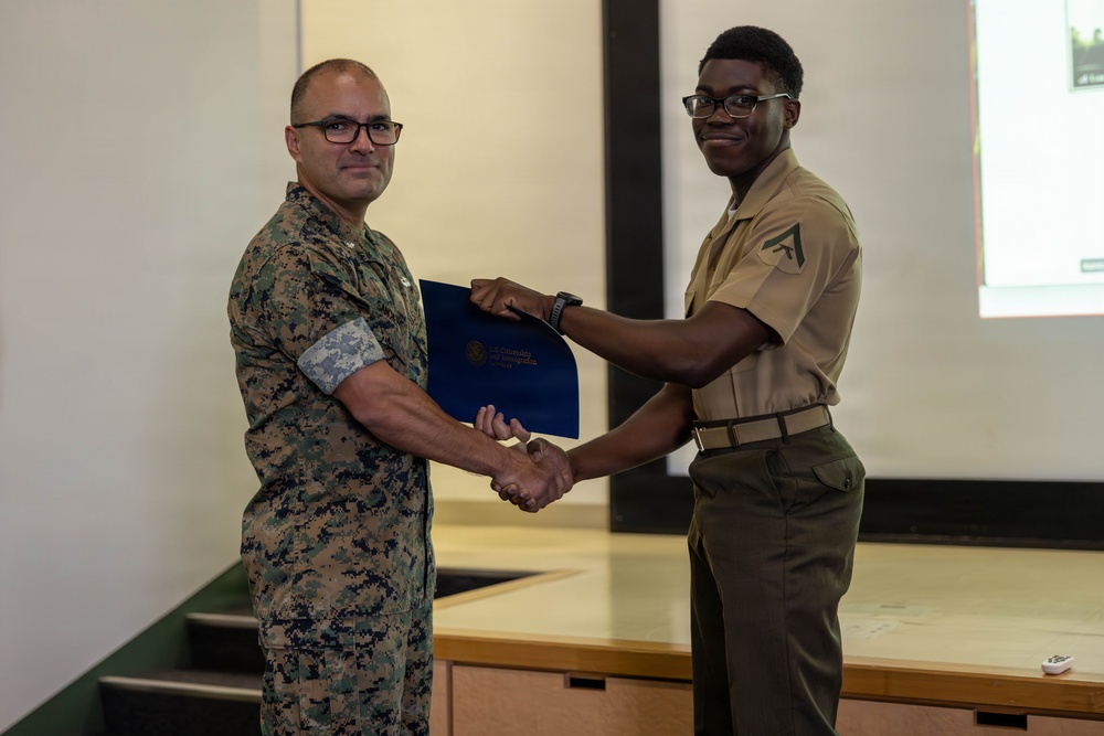 Two military personnel are shaking hands during what appears to be an award or recognition ceremony. One individual in camouflage (MARPAT) uniform is presenting a blue folder or certificate to another in a tan service uniform with lance corporal chevrons. The setting appears to be a briefing room or classroom with a projection screen visible in the background.