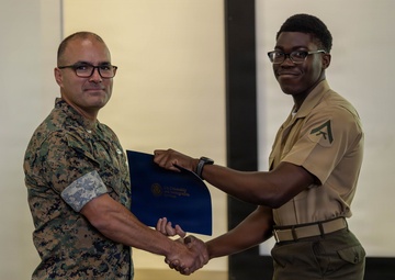 Two military personnel are shaking hands during what appears to be an award or recognition ceremony. One individual in camouflage (MARPAT) uniform is presenting a blue folder or certificate to another in a tan service uniform with lance corporal chevrons. The setting appears to be a briefing room or classroom with a projection screen visible in the background.
