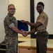 Two military personnel are shaking hands during what appears to be an award or recognition ceremony. One individual in camouflage (MARPAT) uniform is presenting a blue folder or certificate to another in a tan service uniform with lance corporal chevrons. The setting appears to be a briefing room or classroom with a projection screen visible in the background.