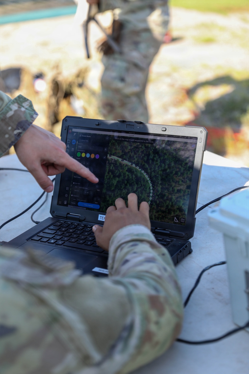 A soldier in camouflage uniform is operating a ruggedized military laptop displaying a drone route-planning software interface with an aerial map view. A dotted circular flight path is visible on the screen over a forested area, and a 'Create Route' panel with color-coded waypoint options is open. A second soldier in camouflage is visible in the blurred background.