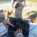 A soldier in camouflage uniform is operating a ruggedized military laptop displaying a drone route-planning software interface with an aerial map view. A dotted circular flight path is visible on the screen over a forested area, and a 'Create Route' panel with color-coded waypoint options is open. A second soldier in camouflage is visible in the blurred background.