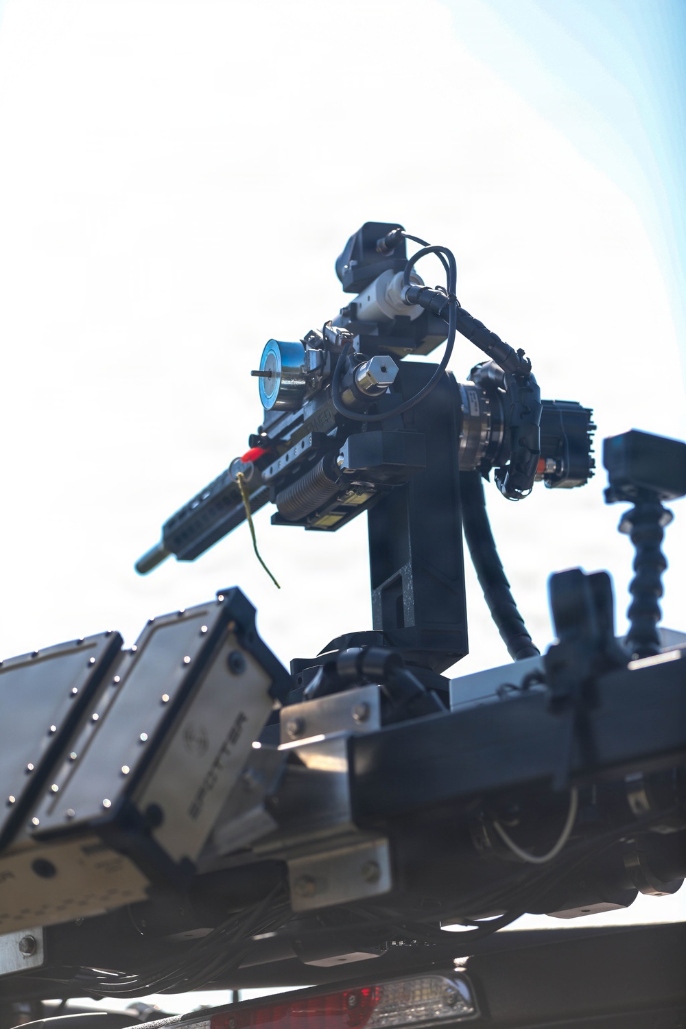 The image shows a close-up of a robotic or remotely operated platform mounted on what appears to be a vehicle or trailer. The system features multiple sensors, cameras, and what appears to be a weapon or tool arm assembly on a pan-tilt mount. The equipment appears to be a tactical or military-grade unmanned ground system photographed against a bright sky.