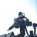 The image shows a close-up of a robotic or remotely operated platform mounted on what appears to be a vehicle or trailer. The system features multiple sensors, cameras, and what appears to be a weapon or tool arm assembly on a pan-tilt mount. The equipment appears to be a tactical or military-grade unmanned ground system photographed against a bright sky.