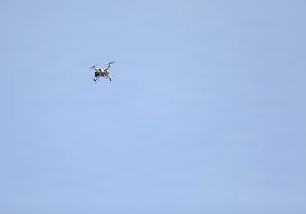 A small quadcopter drone is captured mid-flight against a clear, pale blue sky. The drone appears to be at a moderate altitude with its four rotors visibly spinning. The image is taken from a ground-level perspective looking upward.