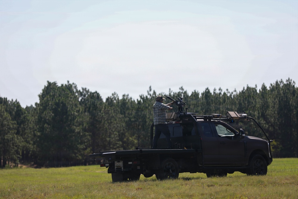 A dark-colored flatbed pickup truck is parked in an open grassy field with a treeline of tall pine trees in the background. A person is standing on or behind the truck operating what appears to be a camera rig or specialized equipment mounted on the vehicle. The scene suggests a film production, wildlife monitoring, or specialized photography operation in a rural outdoor setting.
