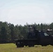 A dark-colored flatbed pickup truck is parked in an open grassy field with a treeline of tall pine trees in the background. A person is standing on or behind the truck operating what appears to be a camera rig or specialized equipment mounted on the vehicle. The scene suggests a film production, wildlife monitoring, or specialized photography operation in a rural outdoor setting.