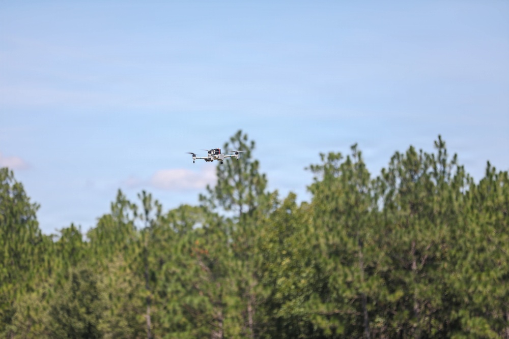 A small quadcopter drone is captured mid-flight against a light blue sky, hovering just above the treeline of a dense forest of tall pine and deciduous trees. The drone appears to be white/light gray with visible rotors and what appears to be a camera or sensor payload mounted underneath. The image has a shallow depth of field with the trees softly blurred in the background.