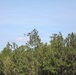 A small quadcopter drone is captured mid-flight against a light blue sky, hovering just above the treeline of a dense forest of tall pine and deciduous trees. The drone appears to be white/light gray with visible rotors and what appears to be a camera or sensor payload mounted underneath. The image has a shallow depth of field with the trees softly blurred in the background.