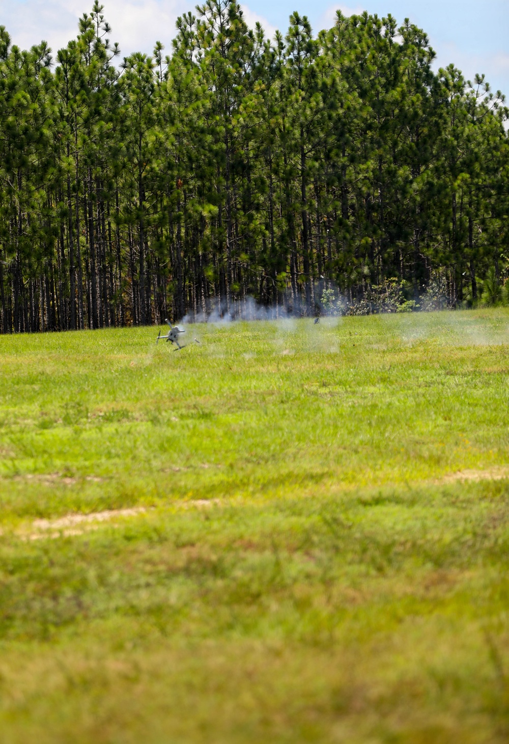 A small unmanned aerial vehicle (UAV/drone) appears to be landing or crashing in an open grassy field, generating a visible smoke trail behind it. The scene is set against a backdrop of tall pine trees under a partly cloudy sky. The smoke and disturbed vegetation suggest a recent impact or controlled landing with propulsion activity.