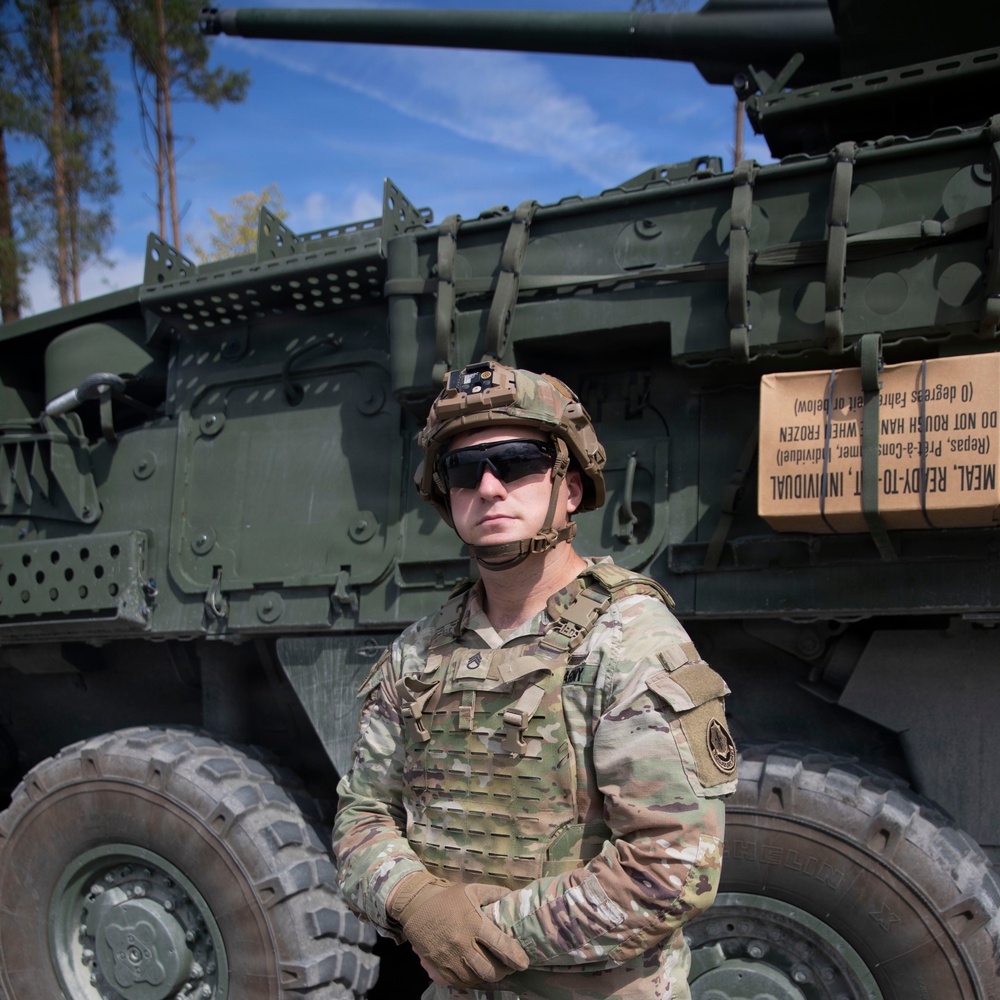 A U.S. Army soldier in full combat gear stands in front of a large wheeled military vehicle, likely a Stryker or similar armored platform, in an outdoor wooded setting. The soldier is wearing Operational Camouflage Pattern (OCP) uniform, a plate carrier vest, combat helmet with mounted device, and tactical sunglasses. A box labeled 'MEAL, READY-TO-EAT, INDIVIDUAL' (MRE) is visible strapped to the vehicle.