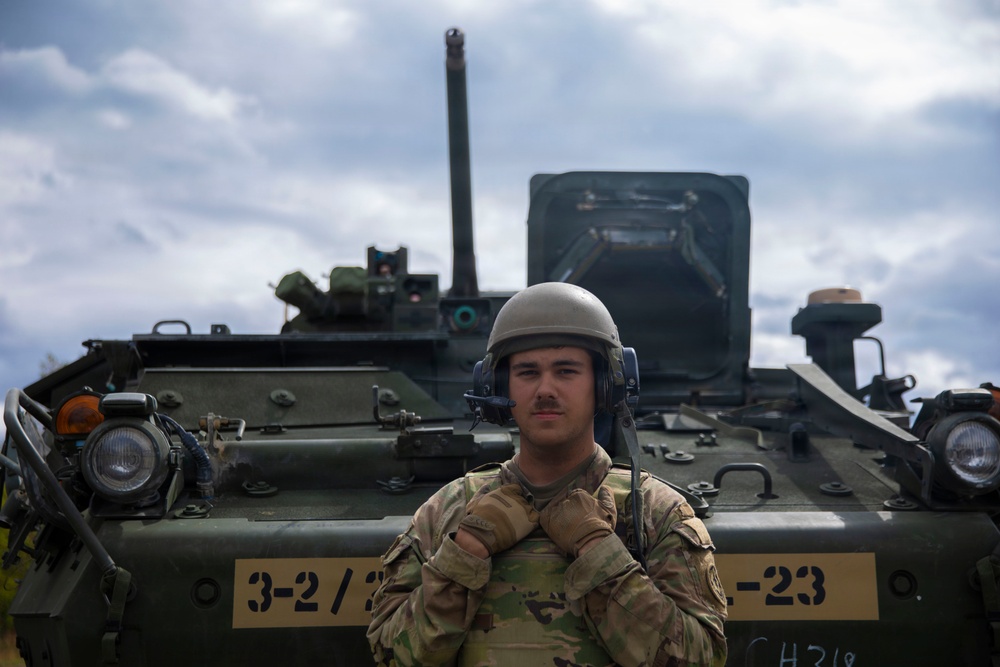 A U.S. Army soldier in MultiCam (OCP) camouflage uniform stands in front of what appears to be a Stryker armored fighting vehicle. The soldier is wearing a combat vehicle crewman (CVC) helmet with integrated communications headset. The vehicle bears the unit marking '3-2/___-23' on its hull.